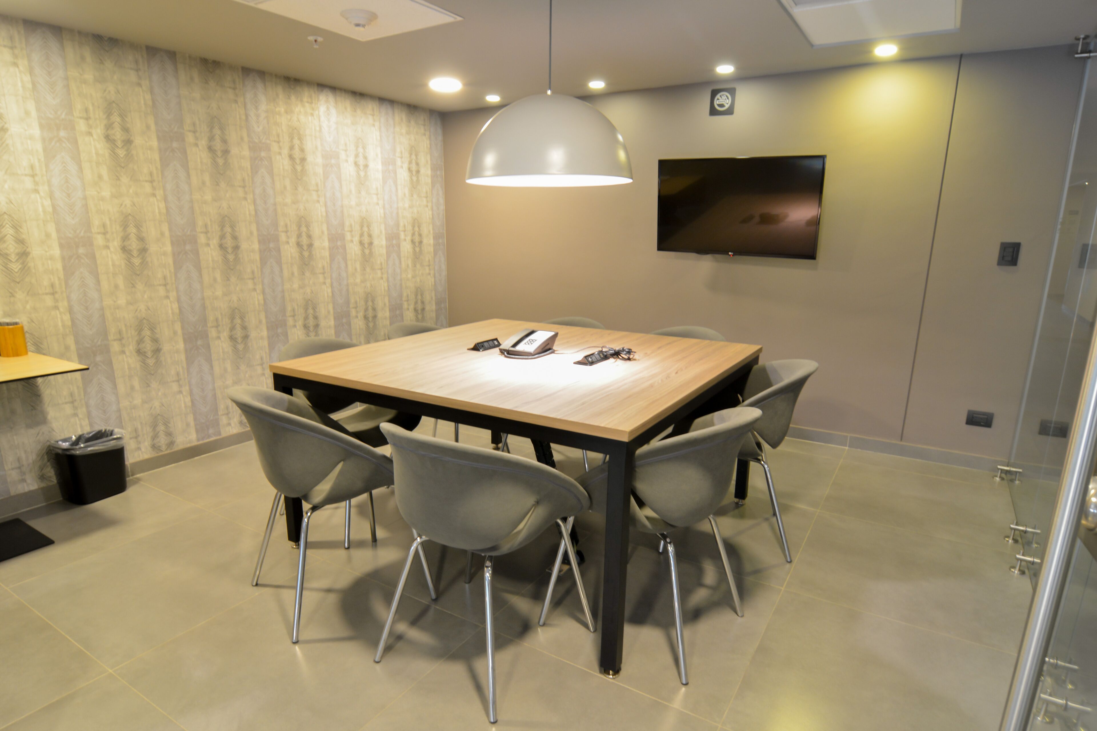 Una sala de conferencias con una mesa de madera, seis sillas, un televisor en la pared y una lámpara en el techo.