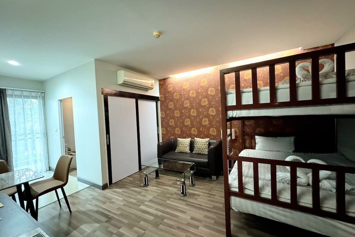 A room with a bunk bed, a sofa, a small table, and a chair. There is a window with curtains, an air conditioning unit, and a patterned wall.