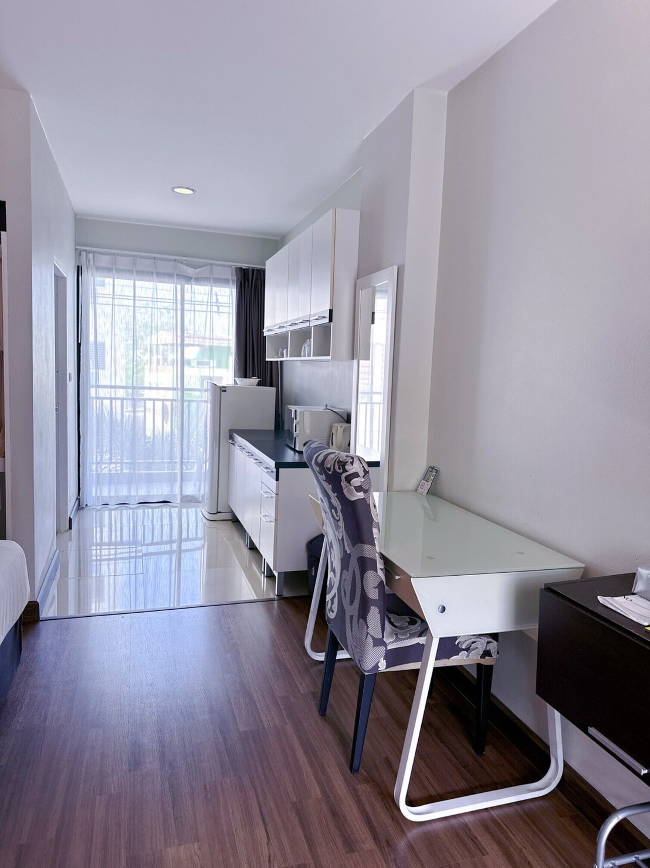 A modern, well-lit room with a glass desk, a chair, and a kitchenette area.