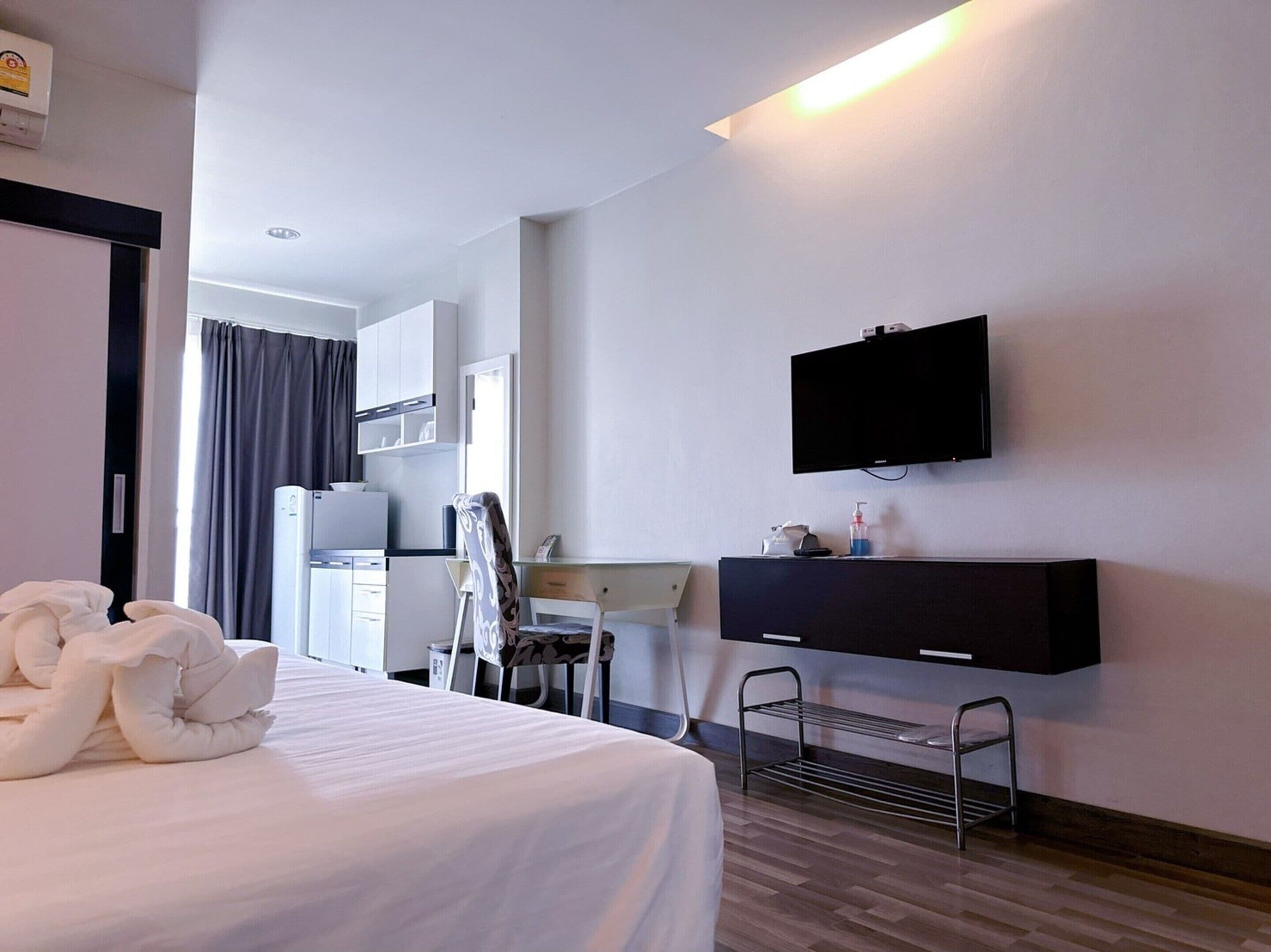 A hotel room with a bed, a desk, a chair, a TV mounted on the wall, and a small shelf with toiletries.