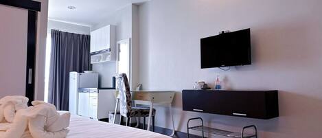 A hotel room with a bed, a desk, a chair, a TV mounted on the wall, and a small shelf with toiletries.