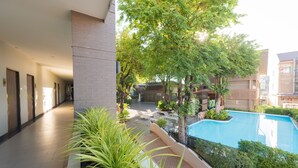 A hotel corridor with a pool area, including a swimming pool, lounge chairs, and potted plants. - Romyen Garden Place (Nakhon Ratchasima)