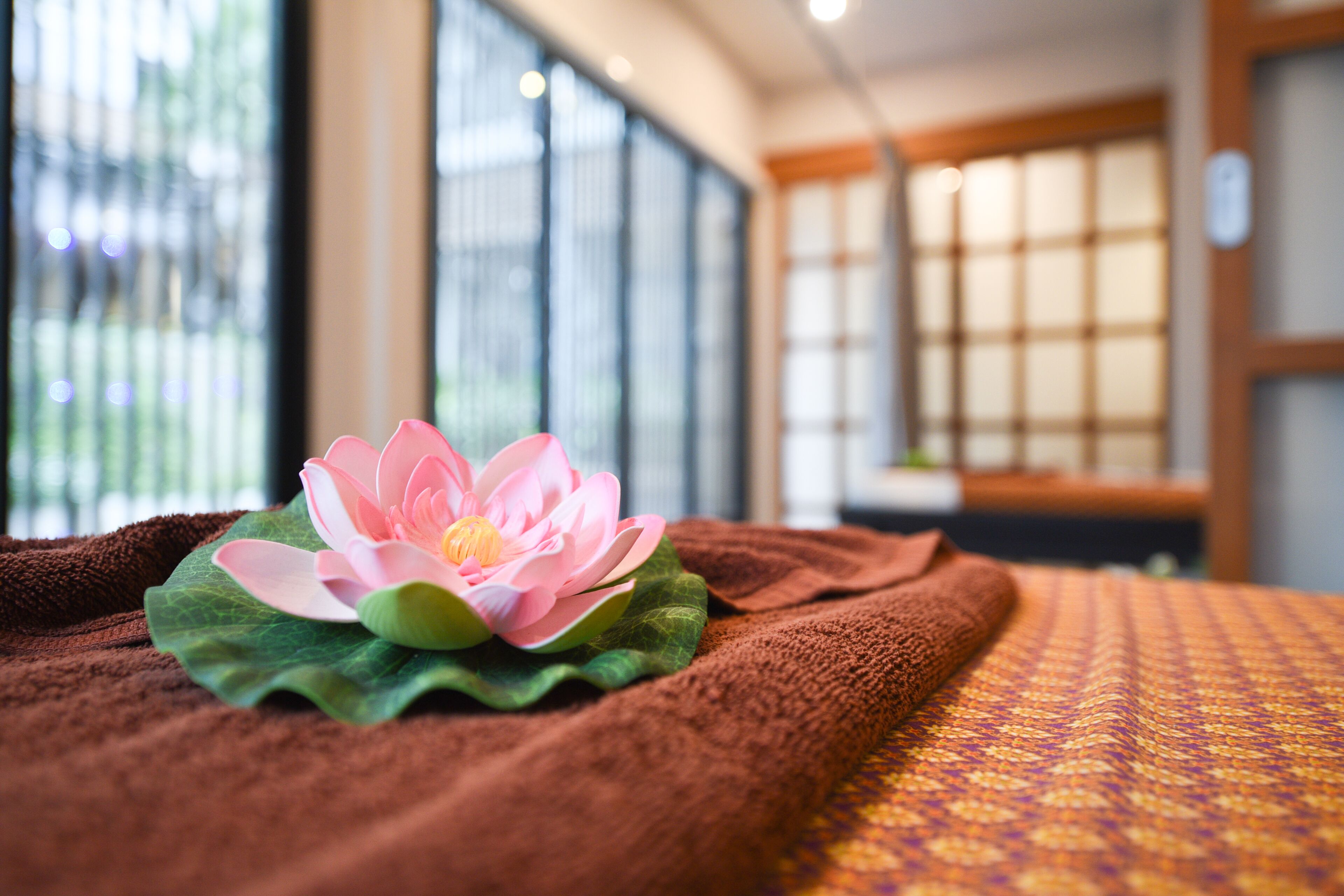 A room with a bed, a lotus flower on a towel, and large windows with a view of greenery.