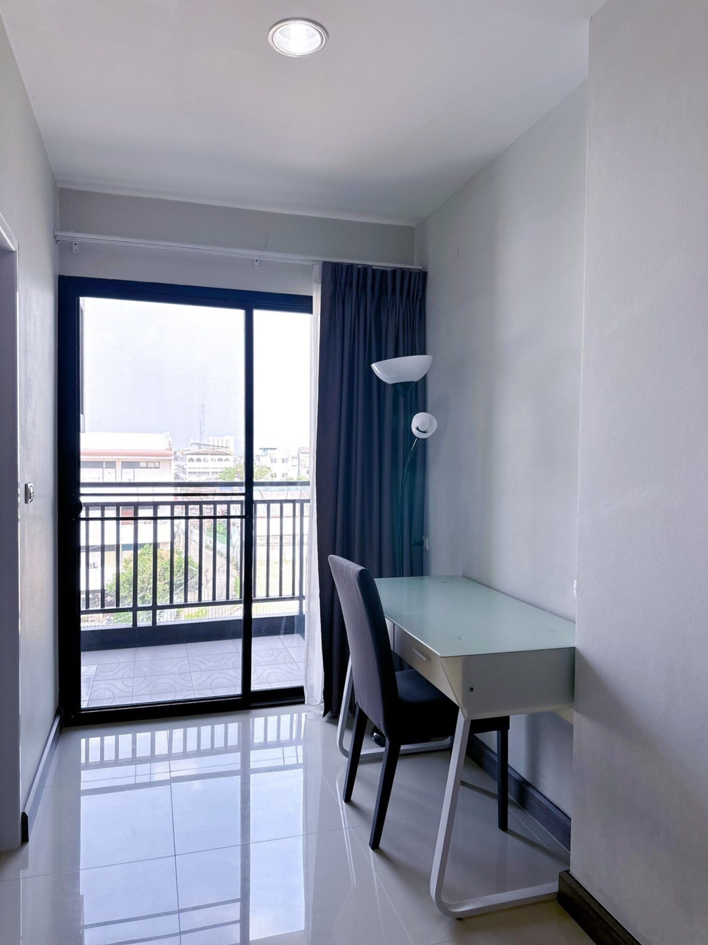 A small, modern room with a desk, chairs, a lamp, and a view of a balcony with a cityscape.