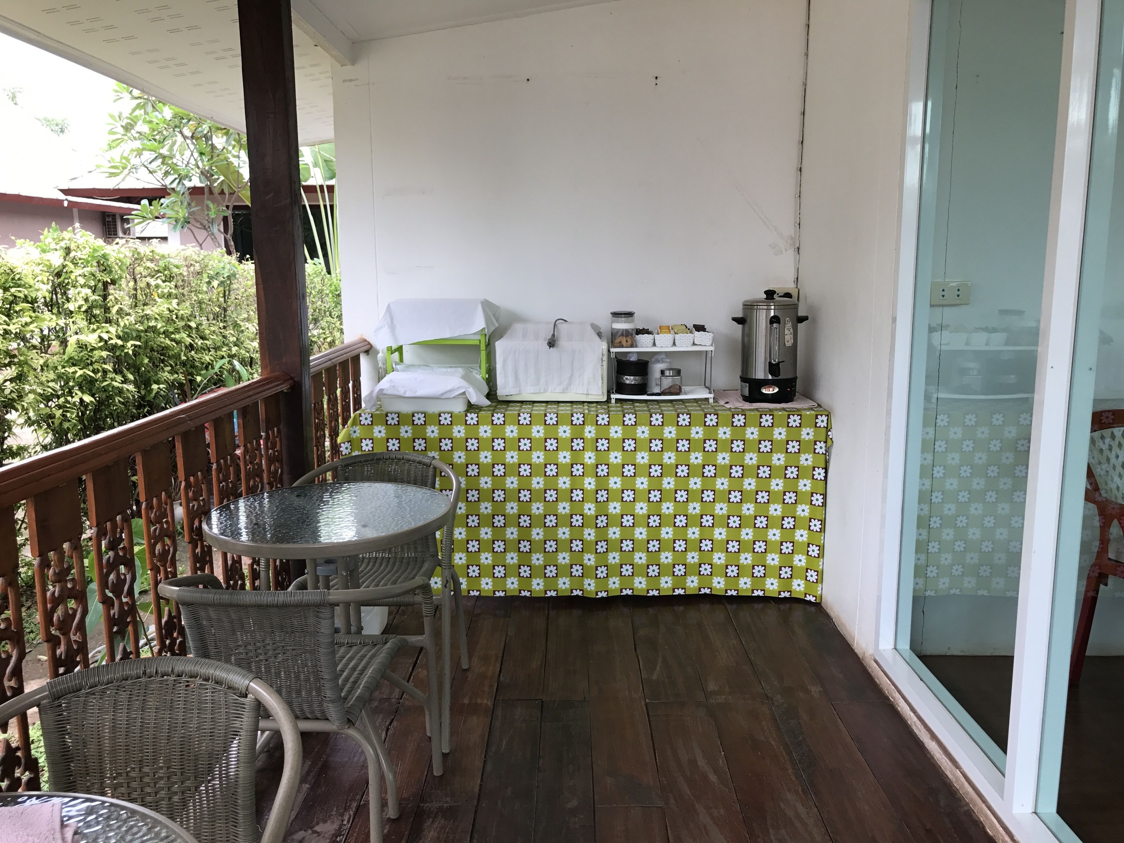 Un balcón con sillas de mimbre, una mesa, mantel con estampado verde, una tetera y un estante con utensilios de cocina.