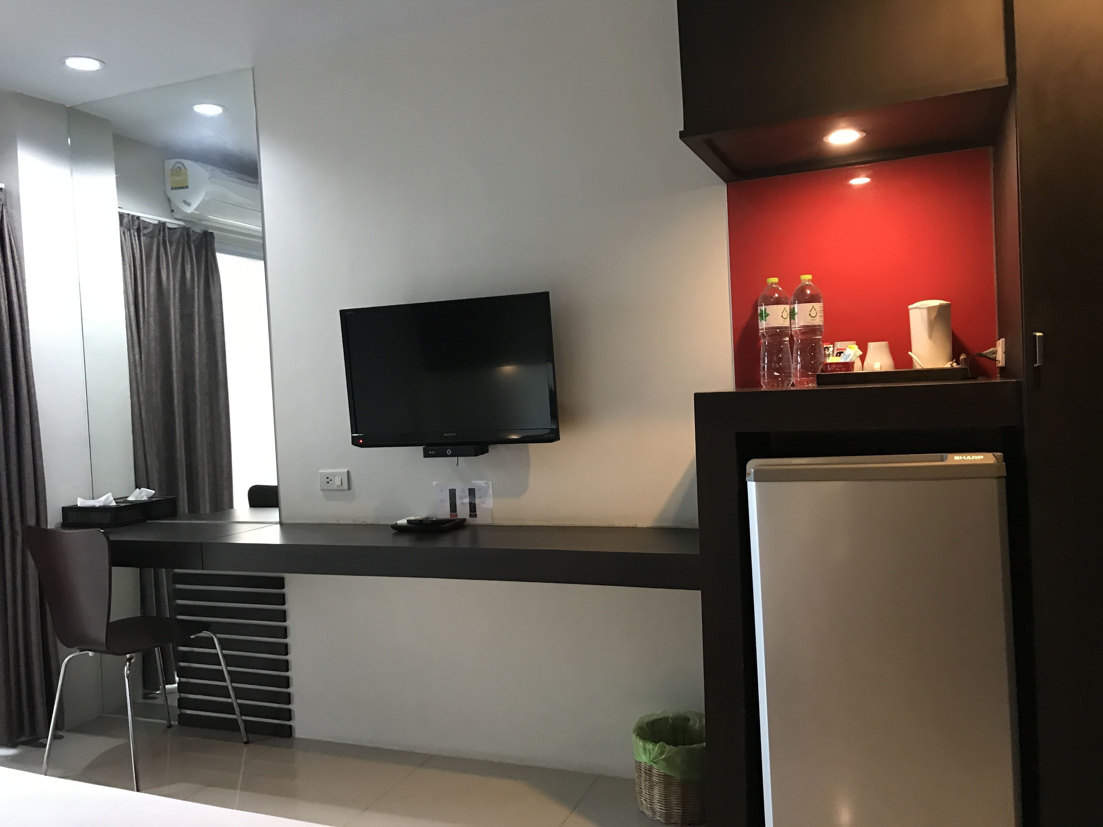 A modern hotel room with a flat-screen TV mounted on the wall, a small kitchenette area with a refrigerator, and a dining table with a chair.
