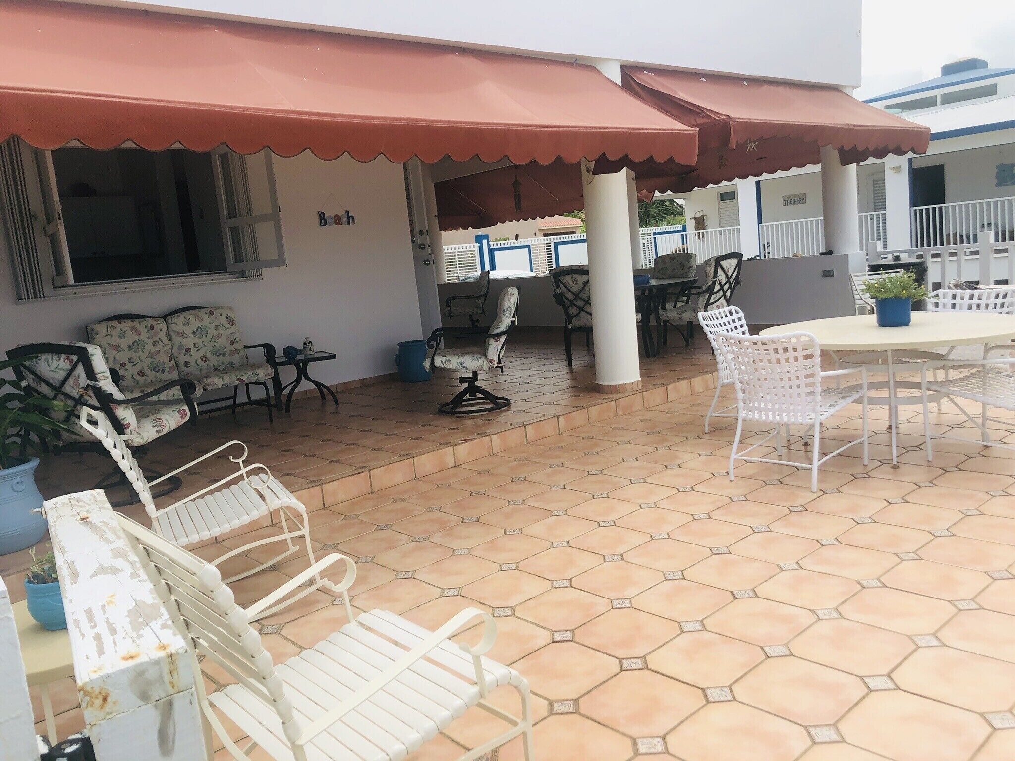 An outdoor patio area with hexagonal tiled flooring, white metal chairs, a round table, and a covered seating area with a red awning.