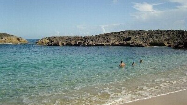Una playa con aguas turquesas cristalinas, afloramientos rocosos y una orilla arenosa.
