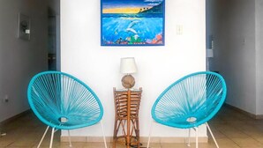 Un pasillo con dos sillas turquesas, una mesita con una lámpara y un cuadro de un paisaje marino en la pared.