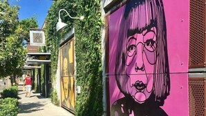 A sidewalk with a mural of a woman with pink hair and polka dots, a street lamp, and a person walking in the distance.