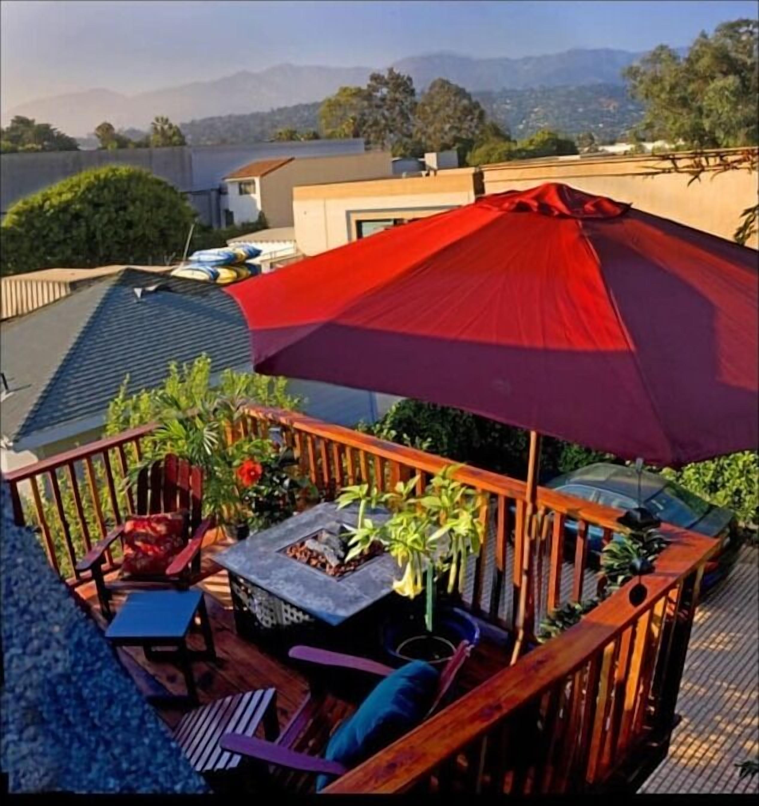 A deck with a red umbrella, wooden furniture, and potted plants.