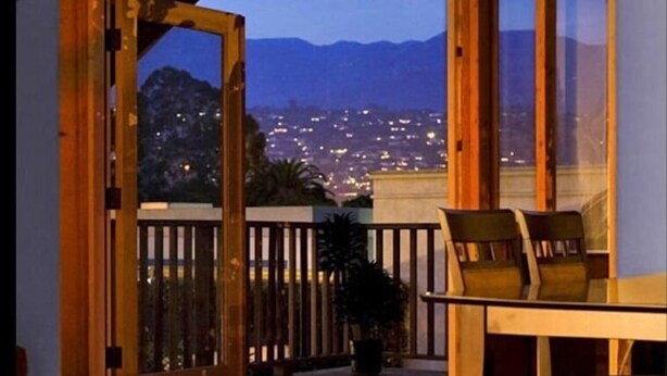 An open balcony with wooden flooring and chairs, overlooking a cityscape at dusk with mountains in the background.