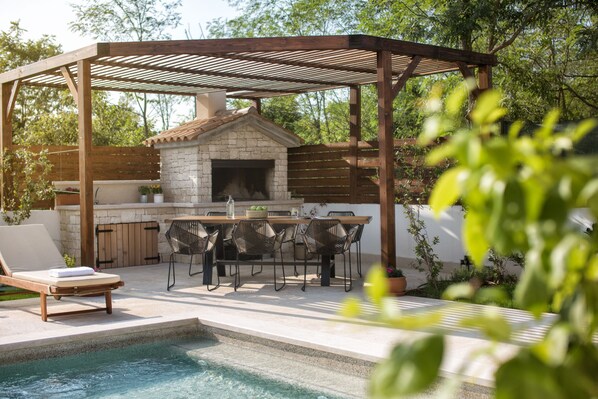 Un patio al aire libre con piscina, una pérgola de madera, chimenea de piedra y zona de comedor con sillas y mesas.