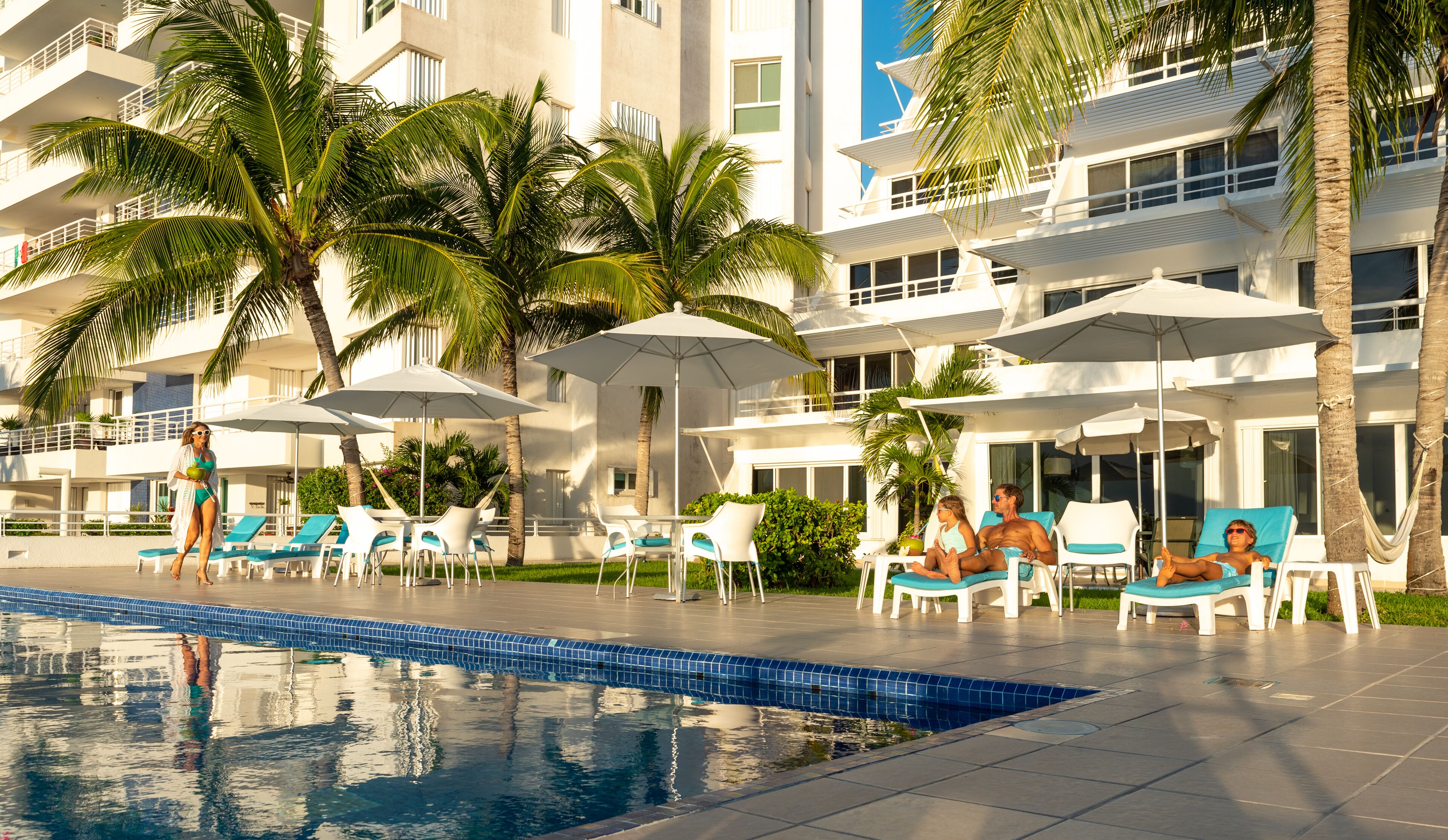 Zona de piscina con sillones de descanso y sombrillas, palmeras y un edificio con balcones.