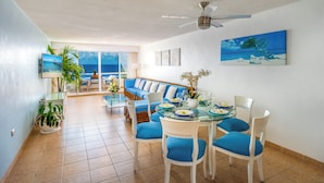 A modern living room with a glass dining table, blue upholstered chairs, a blue sofa, and a large window with a view of the ocean. - Relaxing Oceanfront 2BR Northshore Condo, Awesome snorkeling right in front! (Cozumel)
