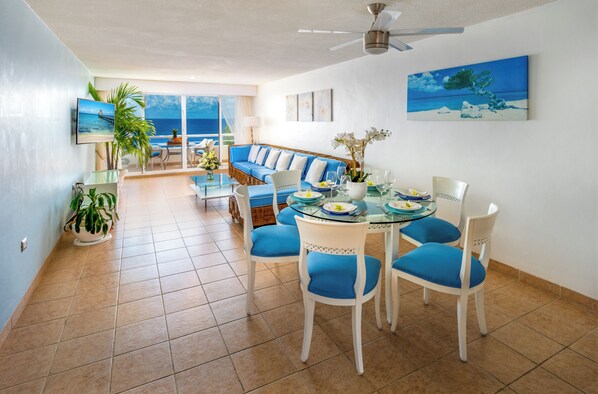 A modern living room with a glass dining table, blue upholstered chairs, a blue sofa, and a large window with a view of the ocean.