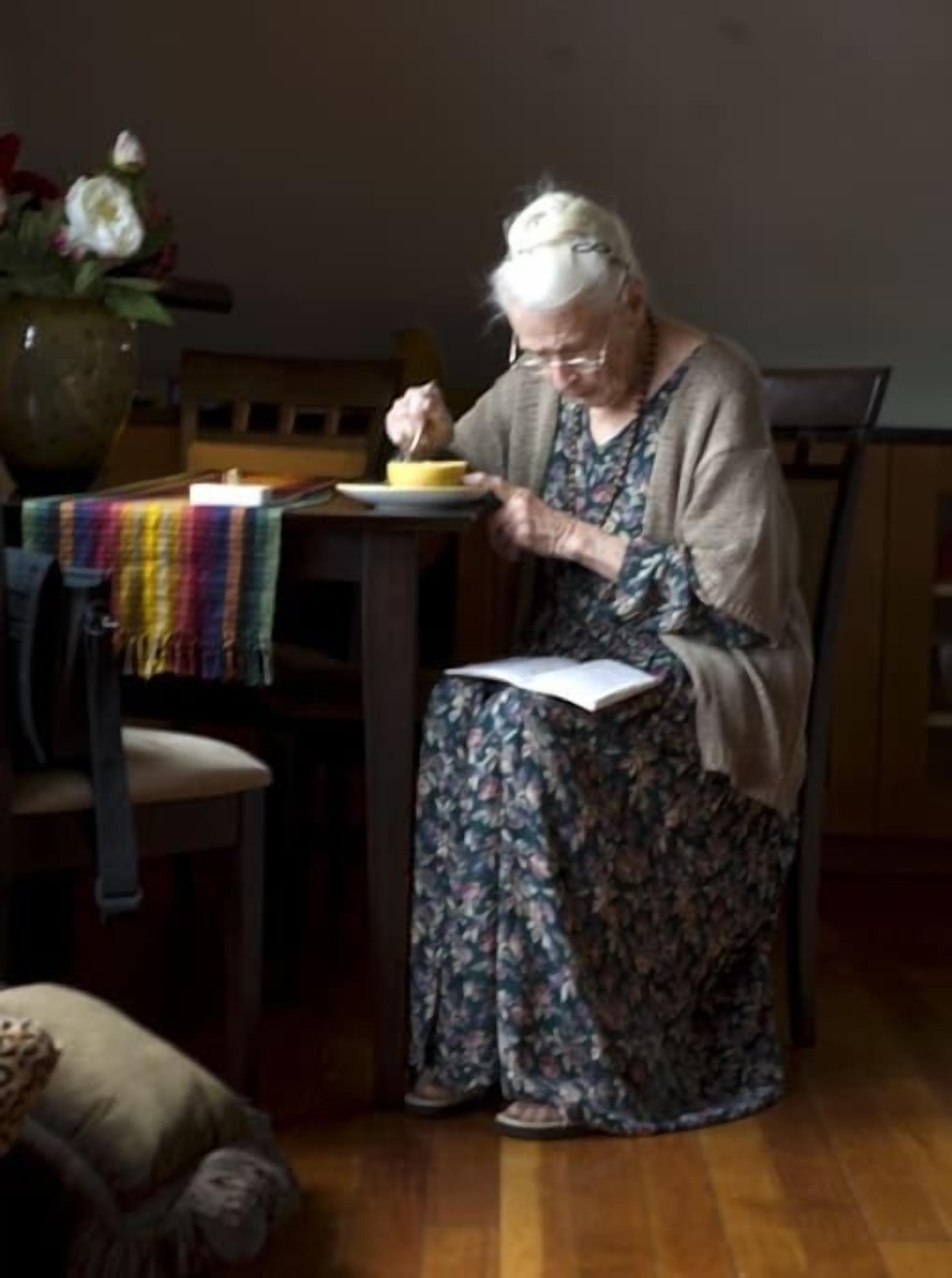Una persona mayor sentada a una mesa con un libro y un plato de comida.
