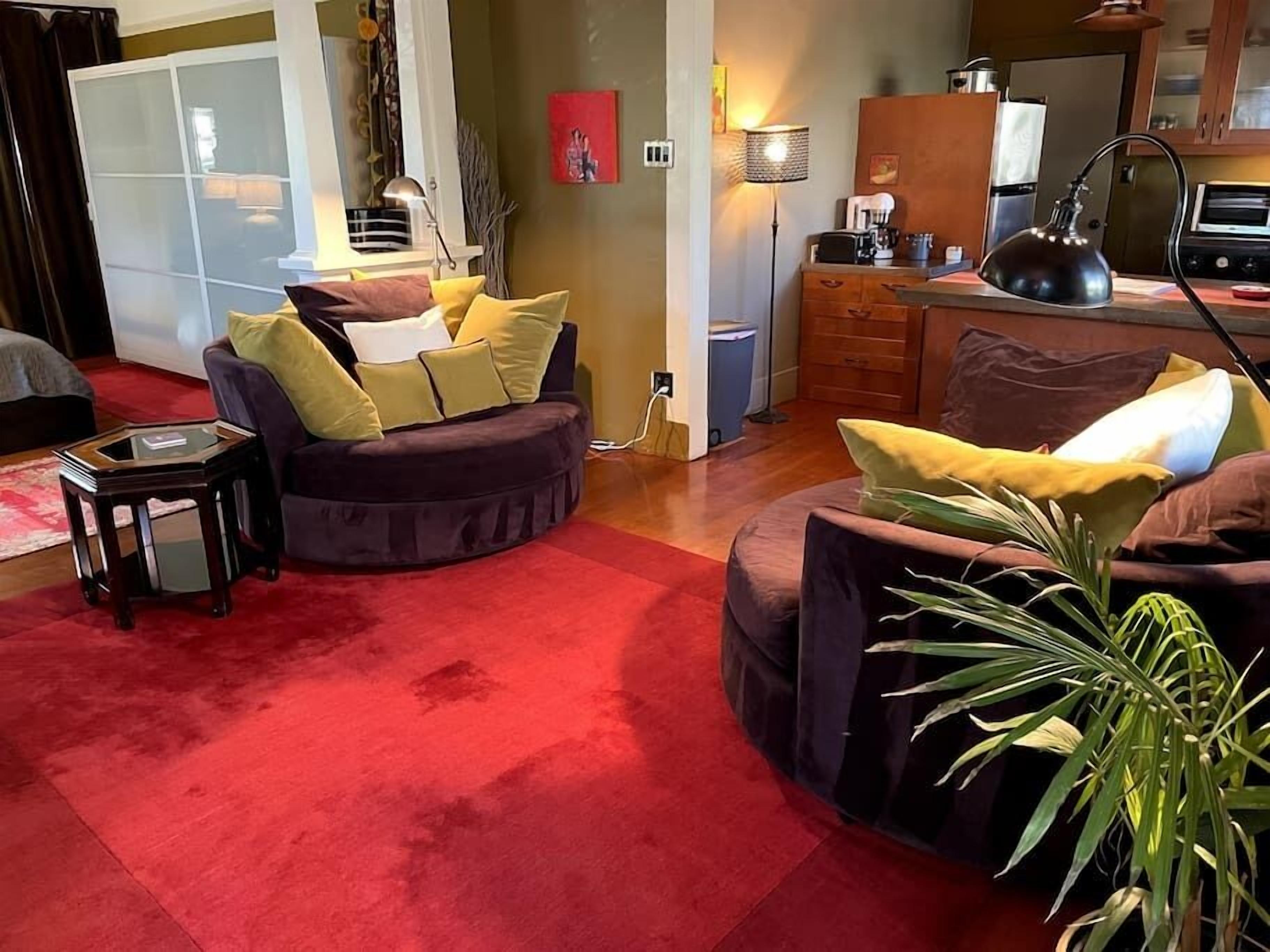 Un salon avec un tapis rouge, deux canapés de couleur foncée ornés de coussins jaunes, une petite table d’appoint en bois, une lampe, et un coin cuisine équipé d’une cuisinière et de placards.