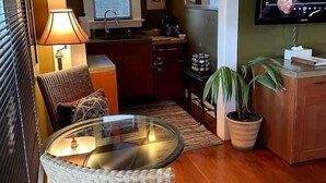 A living room with a glass-top coffee table, wicker chairs, a red rug, a television mounted on the wall, and a kitchen area with a window and a potted plant.