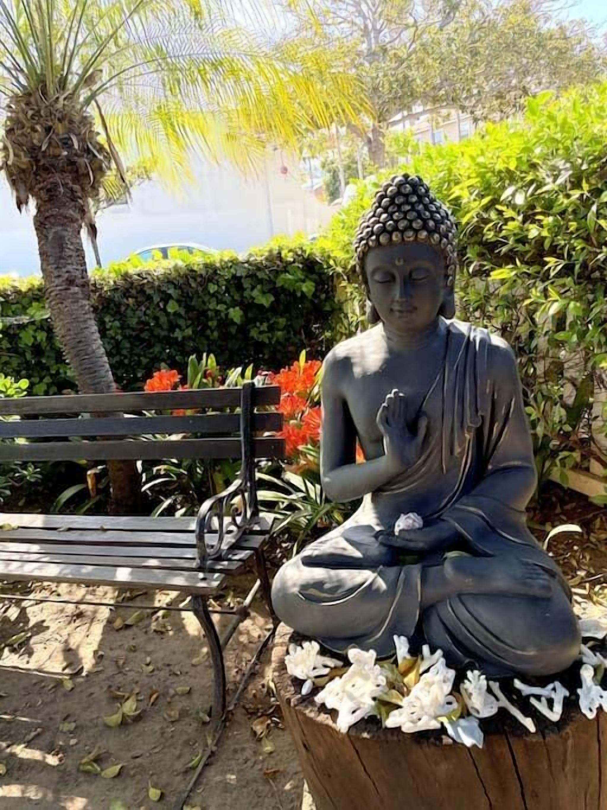 Une statue représentant une figure assise, dans une pose méditative, entourée de verdure et d’un banc.