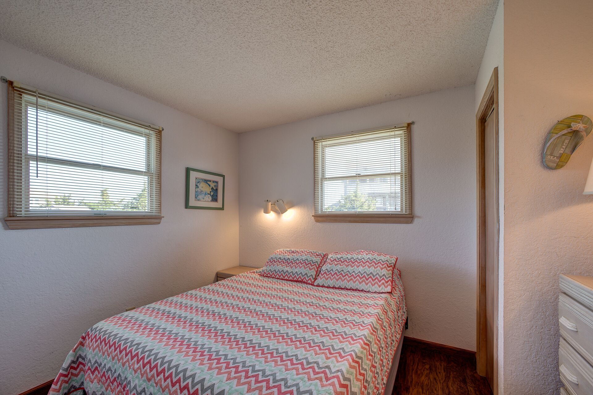 A bedroom with a bed, two windows, a nightstand, a lamp, and a wall-mounted shelf.