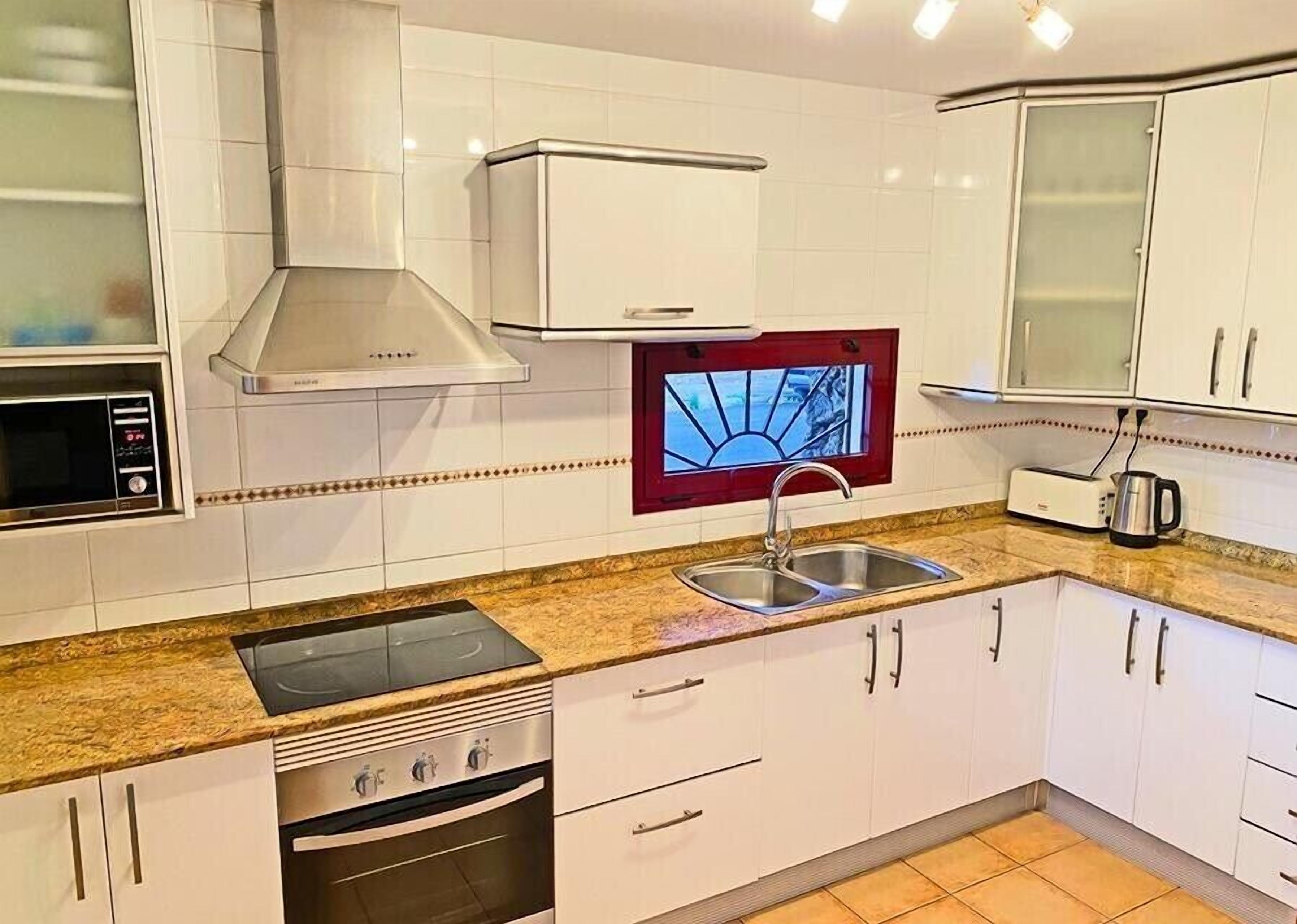 A modern kitchen with white cabinets, a stainless steel sink, a built-in oven, a microwave, and a red-framed window.