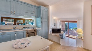 Una cocina con estufa a gas, lavamanos y armarios. Hay una zona de comedor con mesa y sillas. Se ve un balcón con vistas al mar.