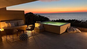 Un moderno espacio lounge al aire libre con sofá, sillones y una bañera exterior con calefacción, con vista a una puesta de sol sobre el mar.