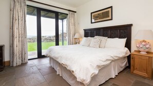 A bedroom with a large bed, a nightstand with a lamp, a painting on the wall, and a view of the outdoors through a glass door. - Traditional & spacious 2 bedroom cottage on the edge of the Lake District  (Selside, Kendal, Cumbria)
