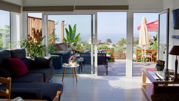 Un soggiorno moderno con divano, tavolino e televisione. Una porta scorrevole in vetro dà accesso a un patio esterno con barbecue e ombrelloni.