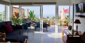 Un salon moderne avec un canapé, une table basse et un téléviseur. Une porte-fenêtre coulissante donne accès à une terrasse extérieure équipée d’un barbecue et de parasols.