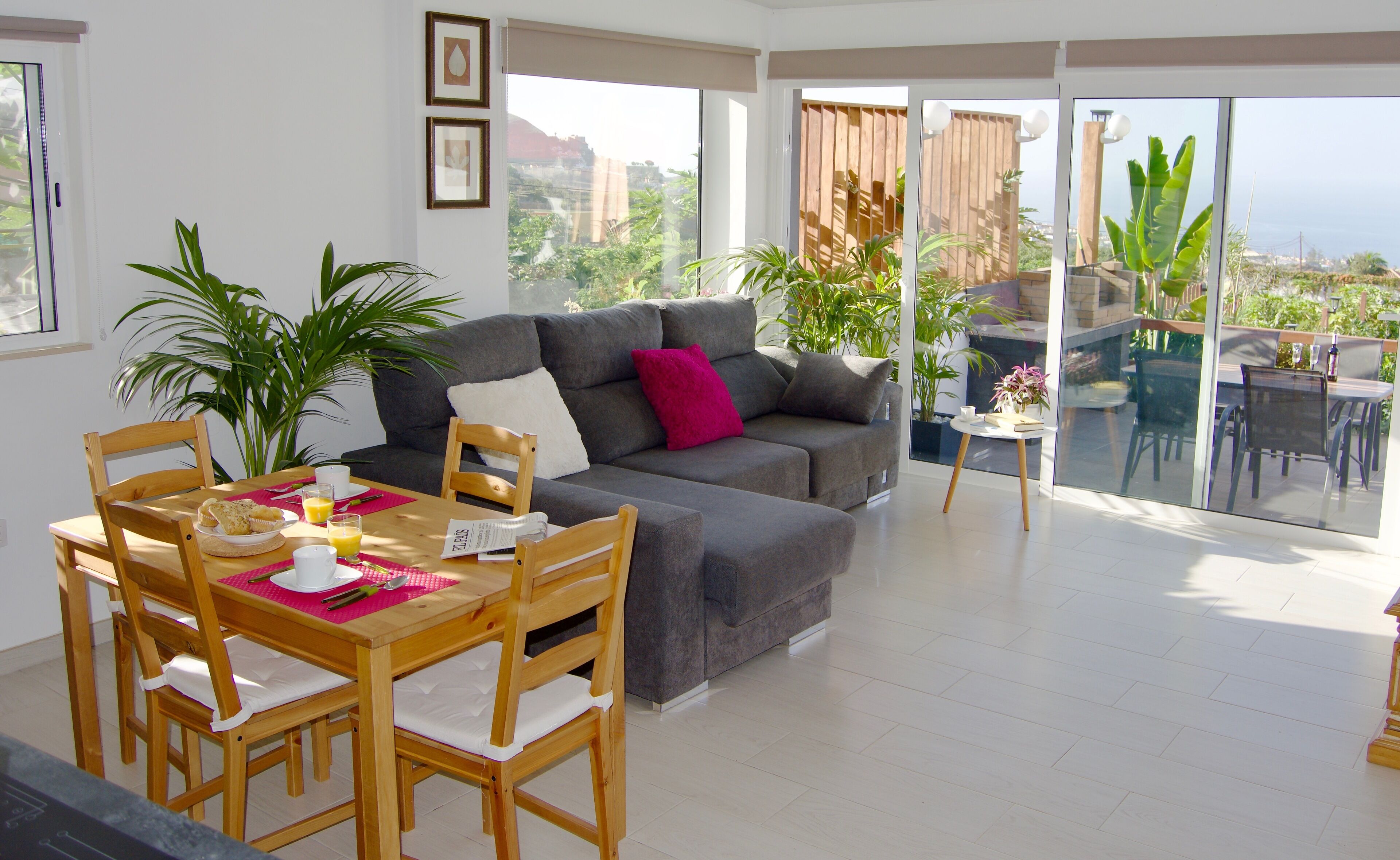 Un salón moderno con un sofá gris, una mesa de comedor de madera y sillas, un balcón con vistas a zonas verdes y edificios.
