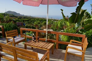 An outdoor seating area with wooden furniture and a pink umbrella, overlooking a scenic landscape with greenery and distant hills.