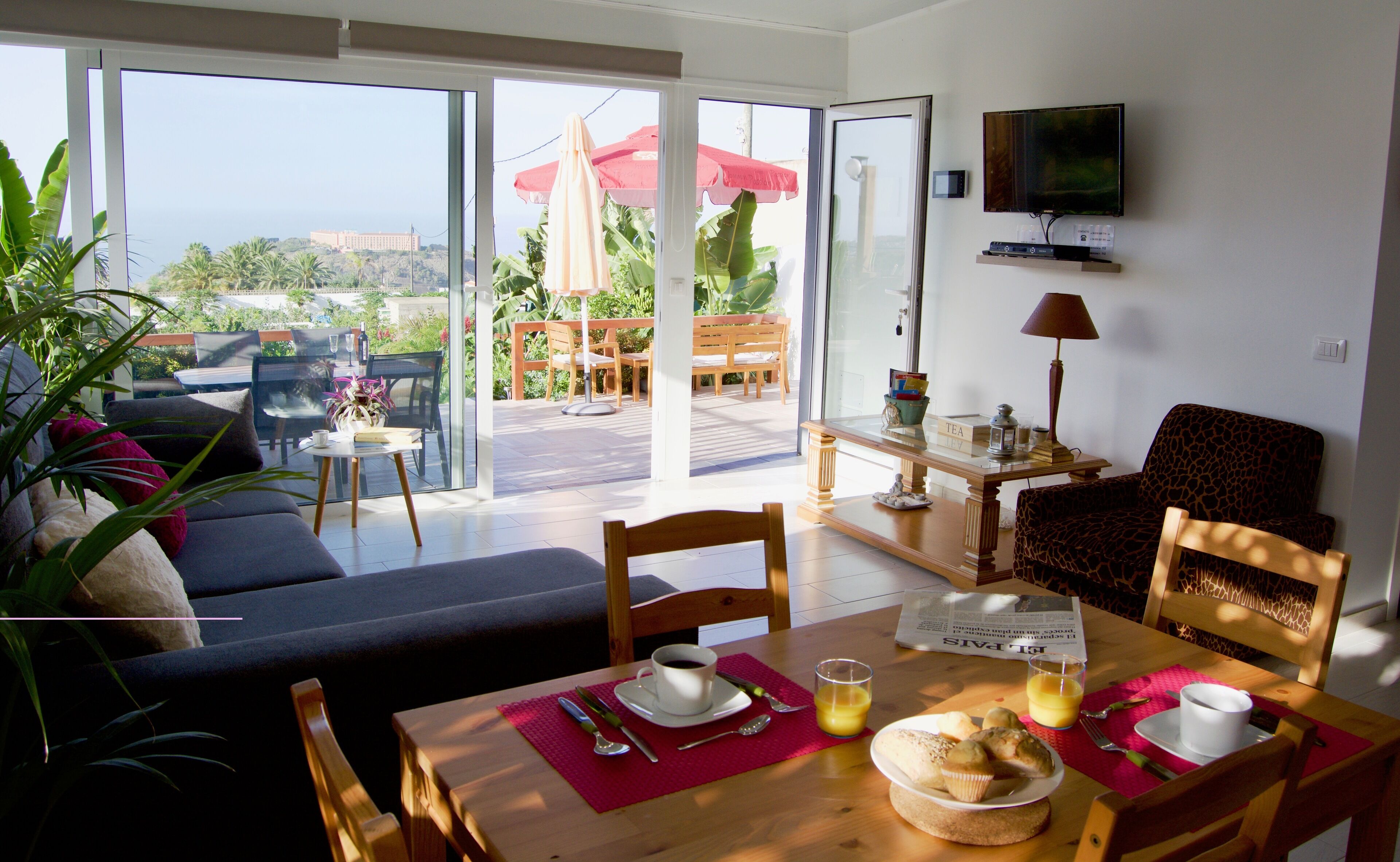A modern living room with a sofa, dining table, and chairs. There is a balcony with outdoor furniture and a TV mounted on the wall.