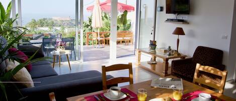A modern living room with a sofa, dining table, and chairs. There is a balcony with outdoor furniture and a TV mounted on the wall.