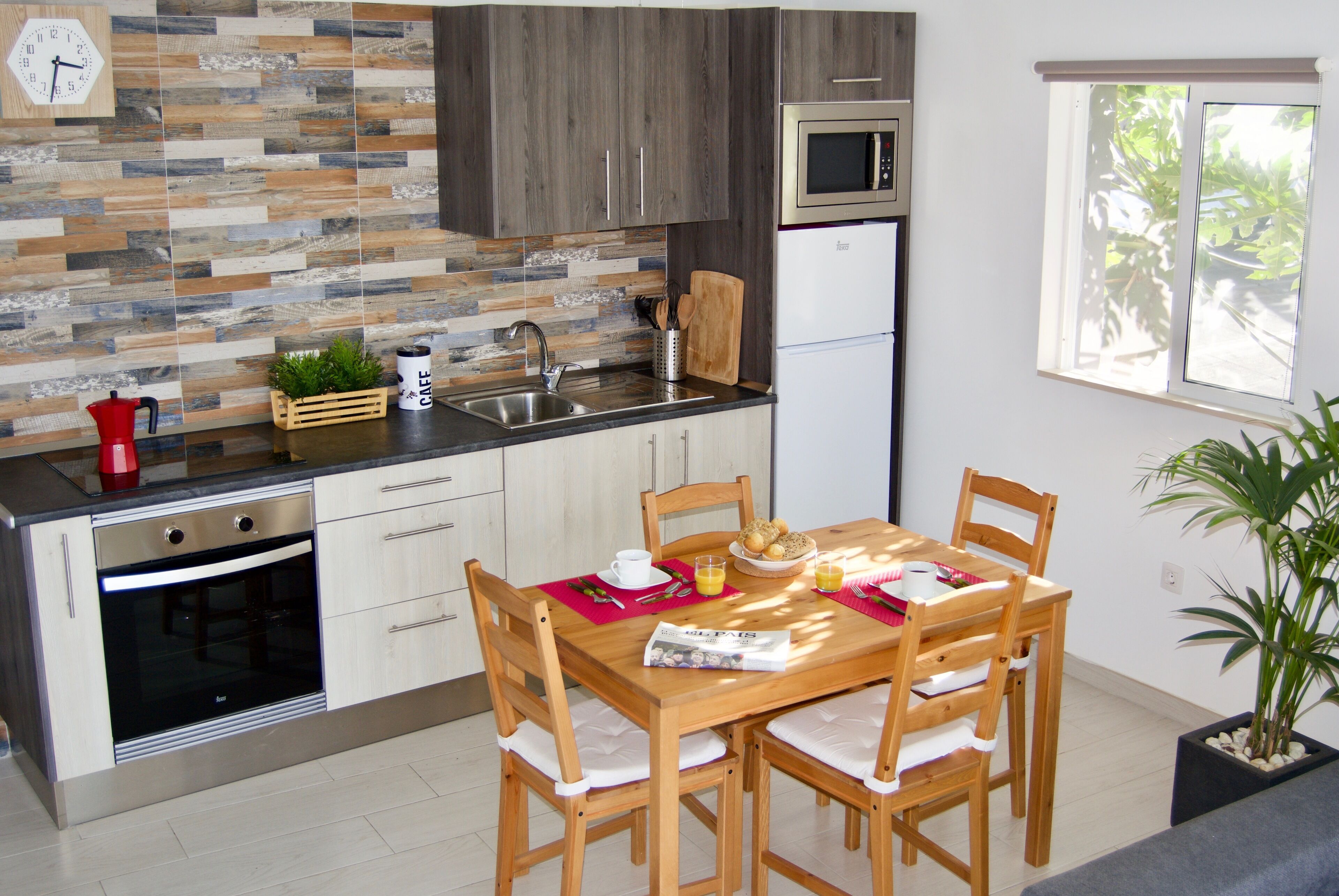 A modern kitchen with a wooden dining table, chairs, and a window with a view of greenery.