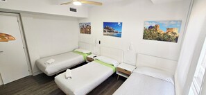 Three single beds with white mattresses and pillows, each with a nightstand and drawer for storage, complemented by a ceiling fan and framed photographs.