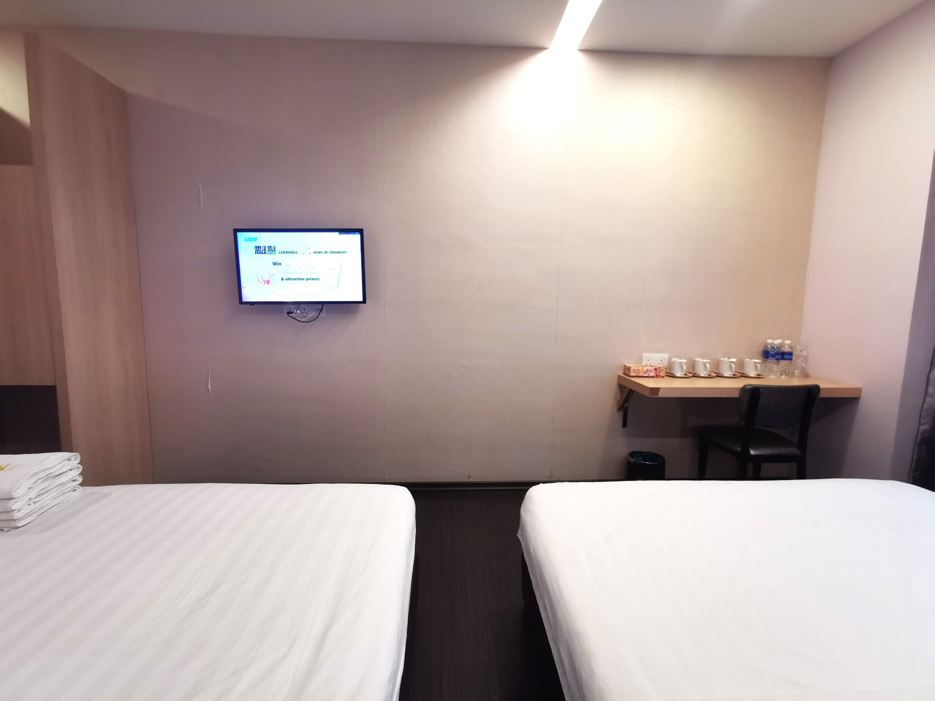 Habitación de hotel con dos camas, un escritorio con silla, un televisor montado en la pared y un cesto de basura pequeño.