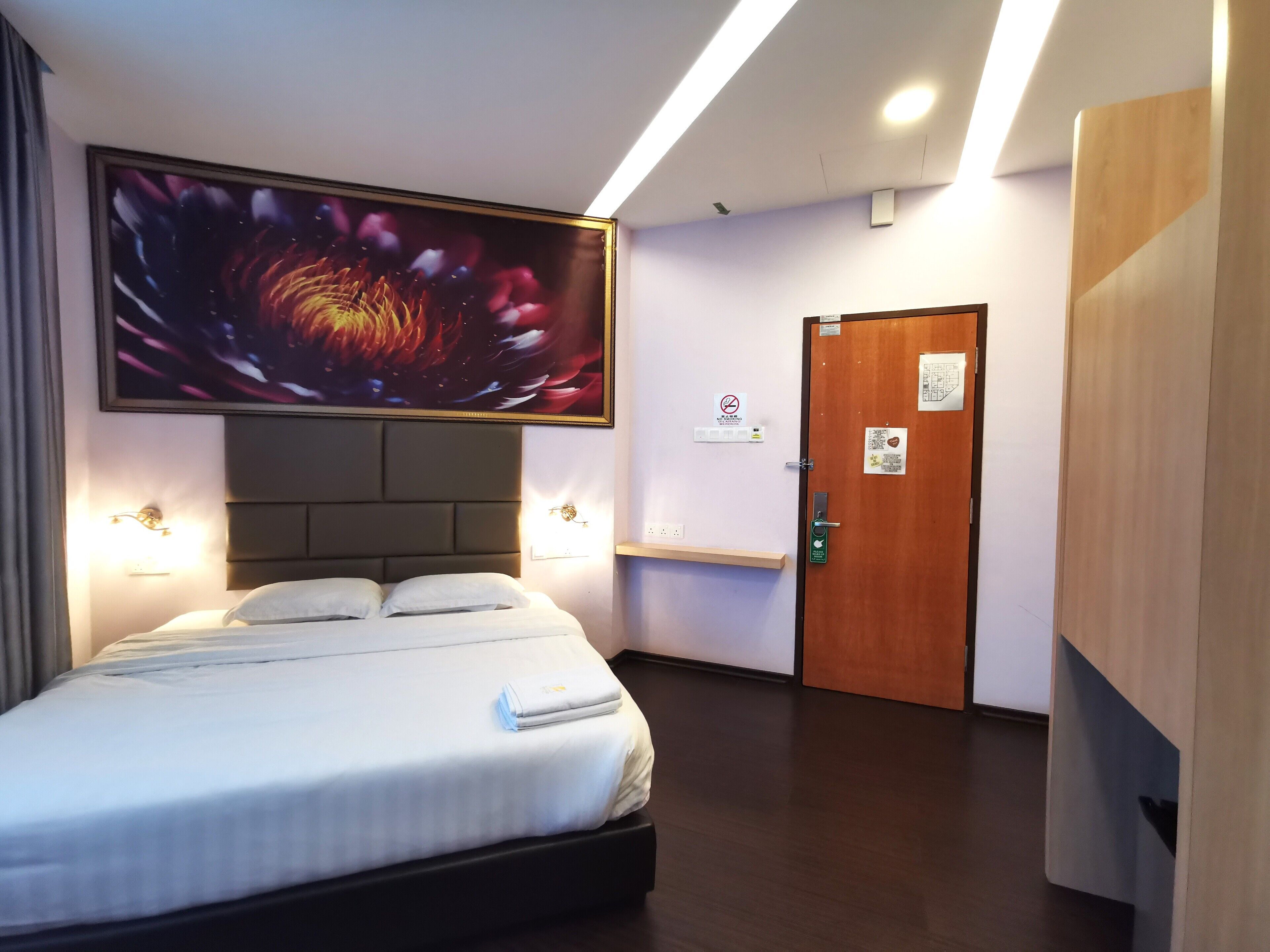 A hotel room with a large bed, a headboard, a wall-mounted TV, a door with a sign, and a wooden wardrobe.