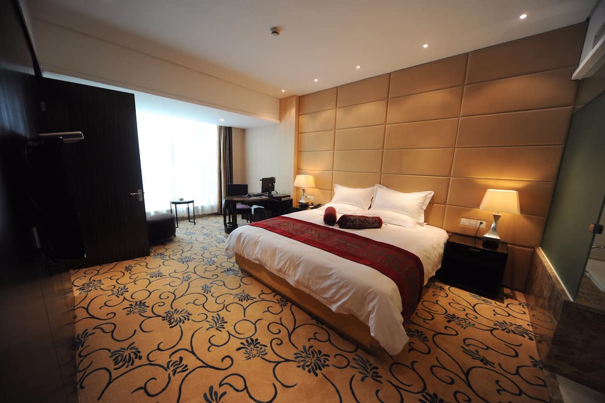 A hotel room with a large bed, two bedside tables with lamps, a desk with a chair, and a large window.