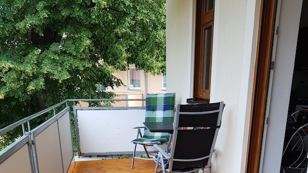 Ett trädäck med ett bord och stolar, ett metallräcke och en dörr som leder in till ett utrymme inomhus.