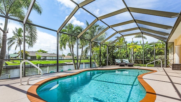An indoor swimming pool with a curved edge, surrounded by a covered area with metal beams and a view of palm trees and a body of water. - Dream villa with tropical garden on the canal (Cape Coral)