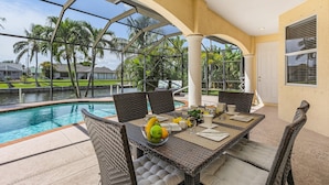 A poolside dining area with wicker furniture, a glass-enclosed patio, and a view of a swimming pool and palm trees. - Dream villa with tropical garden on the canal (Cape Coral)