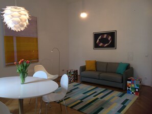 A modern living room with a round white table, white chairs, a gray sofa with colorful cushions, a multicolored rug, and a hanging light fixture. - Absolute quiet location with balcony and still in a few minutes in the center (wien)