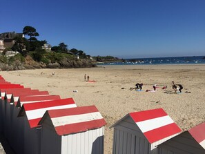 Une plage avec des cabanes de plage rayées de rouge et de blanc, un rivage sablonneux et des bateaux sur l’eau.