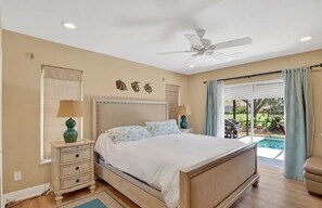 A bedroom with a bed, two bedside tables, a ceiling fan, a window with curtains, and a view of an outdoor area.