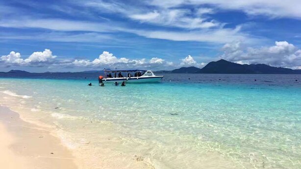 A beach with clear turquoise water, a boat with people on it, and mountains in the background. - Cube Bed Station - Hostel (Semporna)