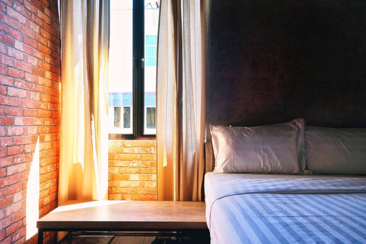 A hotel room with a brick wall, a window with curtains, a bed with pillows, and a wooden table.