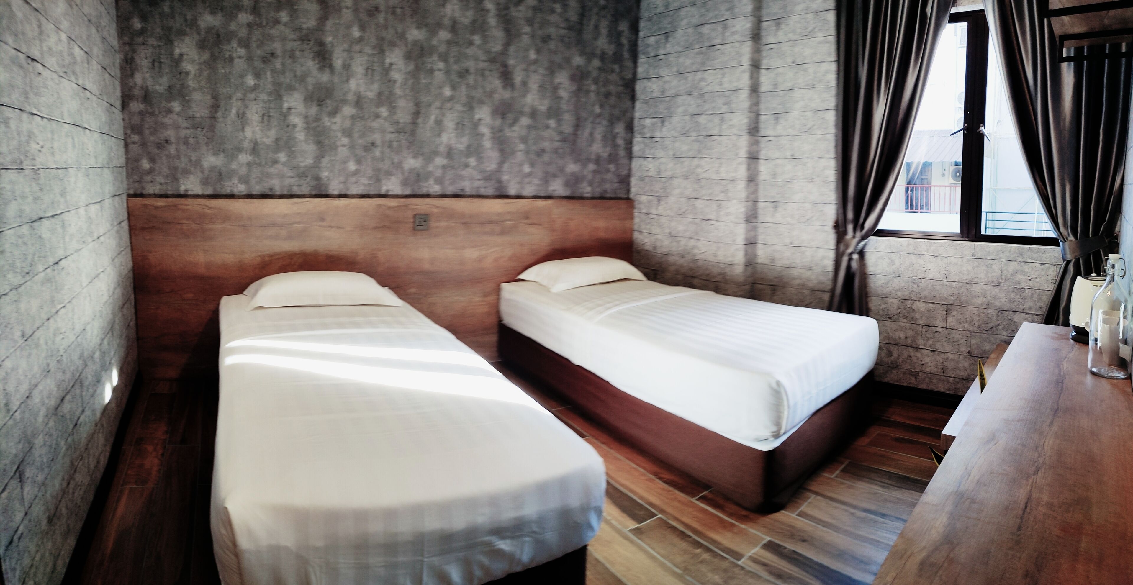 Una habitación con dos camas individuales, un cabecero de madera, una mesita pequeña y una ventana con cortinas.