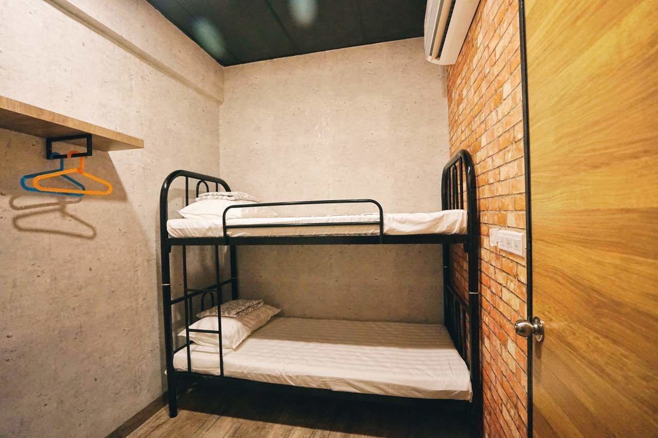 Habitación con literas, pared de ladrillo, puerta de madera y un estante con perchas.